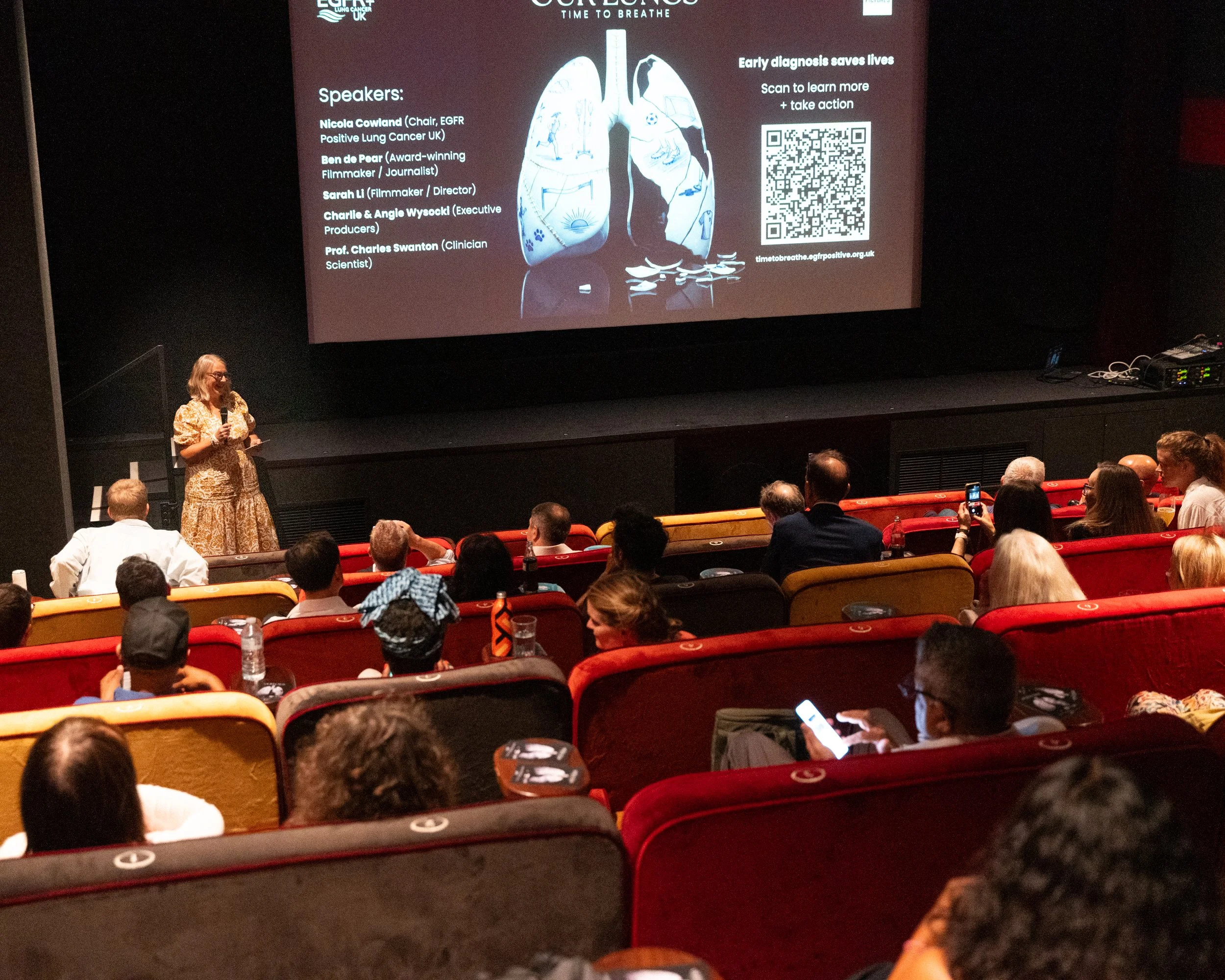 A woman is standing on stage in front of an audience, giving a presentation about lung health. There is a large screen displaying a diagram of human lungs, sponsors' logos, and information about early diagnosis saving lives, along with a QR code.