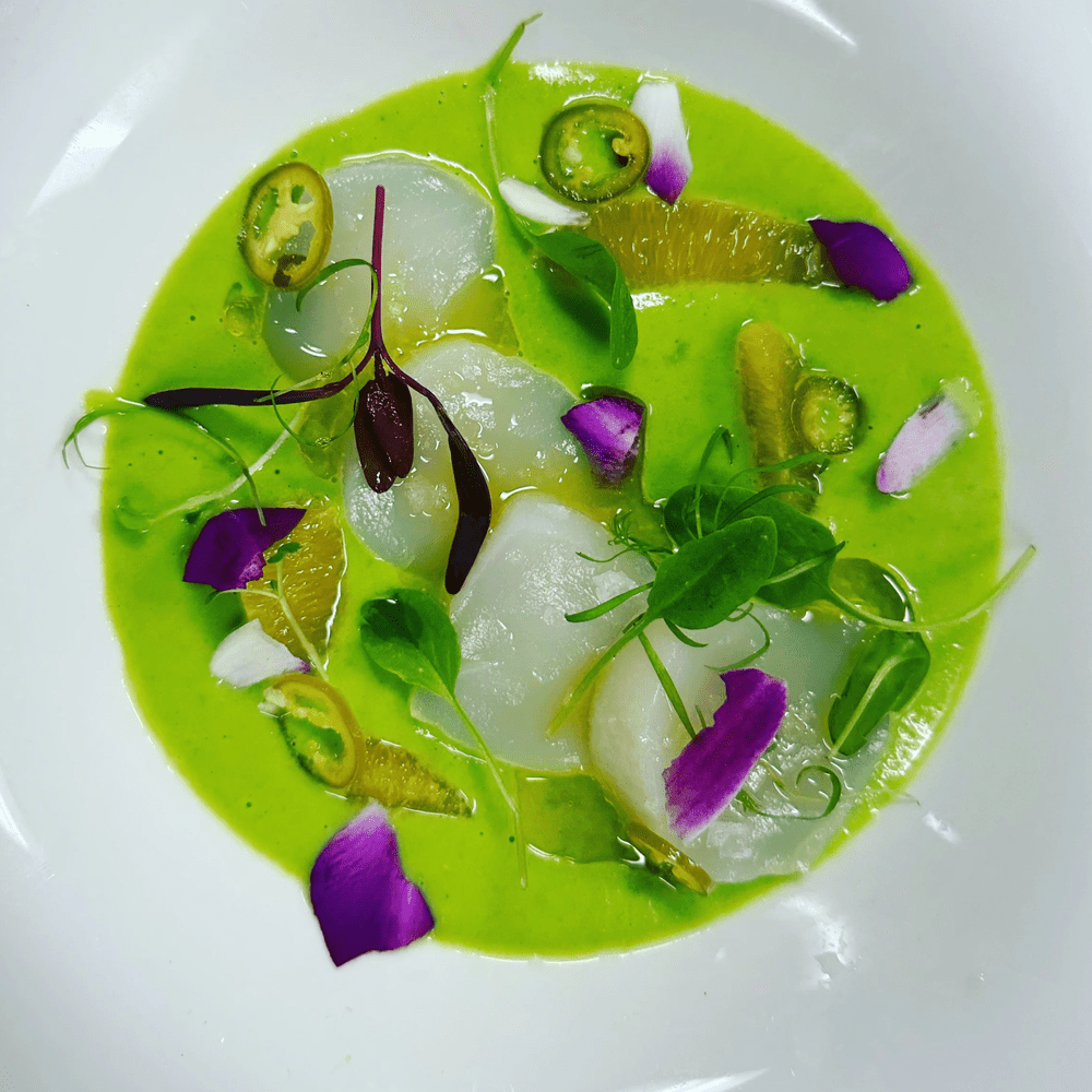 Delicately sliced scallops in a vibrant green sauce with citrus, jalapeño, herbs and edible flowers, inspired by refined culinary experiences.