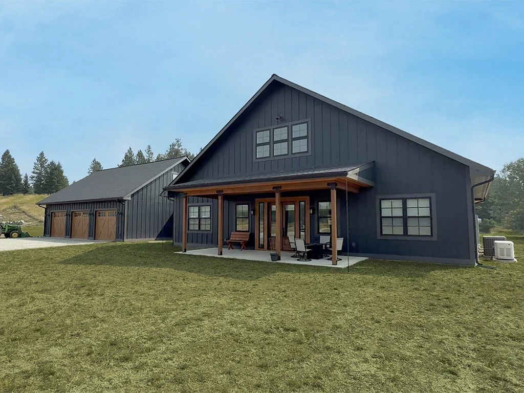 A modern dark blue house with a three-car garage and wooden accents, set on a grassy field under a cloudy sky with trees in the background.