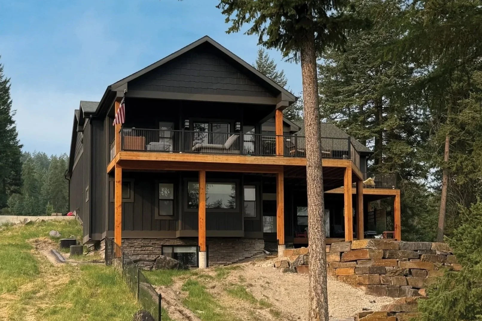 A modern two-story house with outdoor balcony, black siding, surrounded by trees, with a bright blue sky in the background.