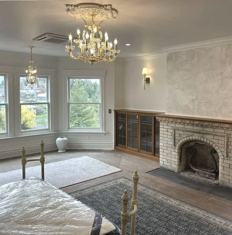 Bedroom with large bay windows, a chandelier, a fireplace with a brick surround, wall sconces, and a built-in wooden cabinet, with a bed partly visible in the foreground.