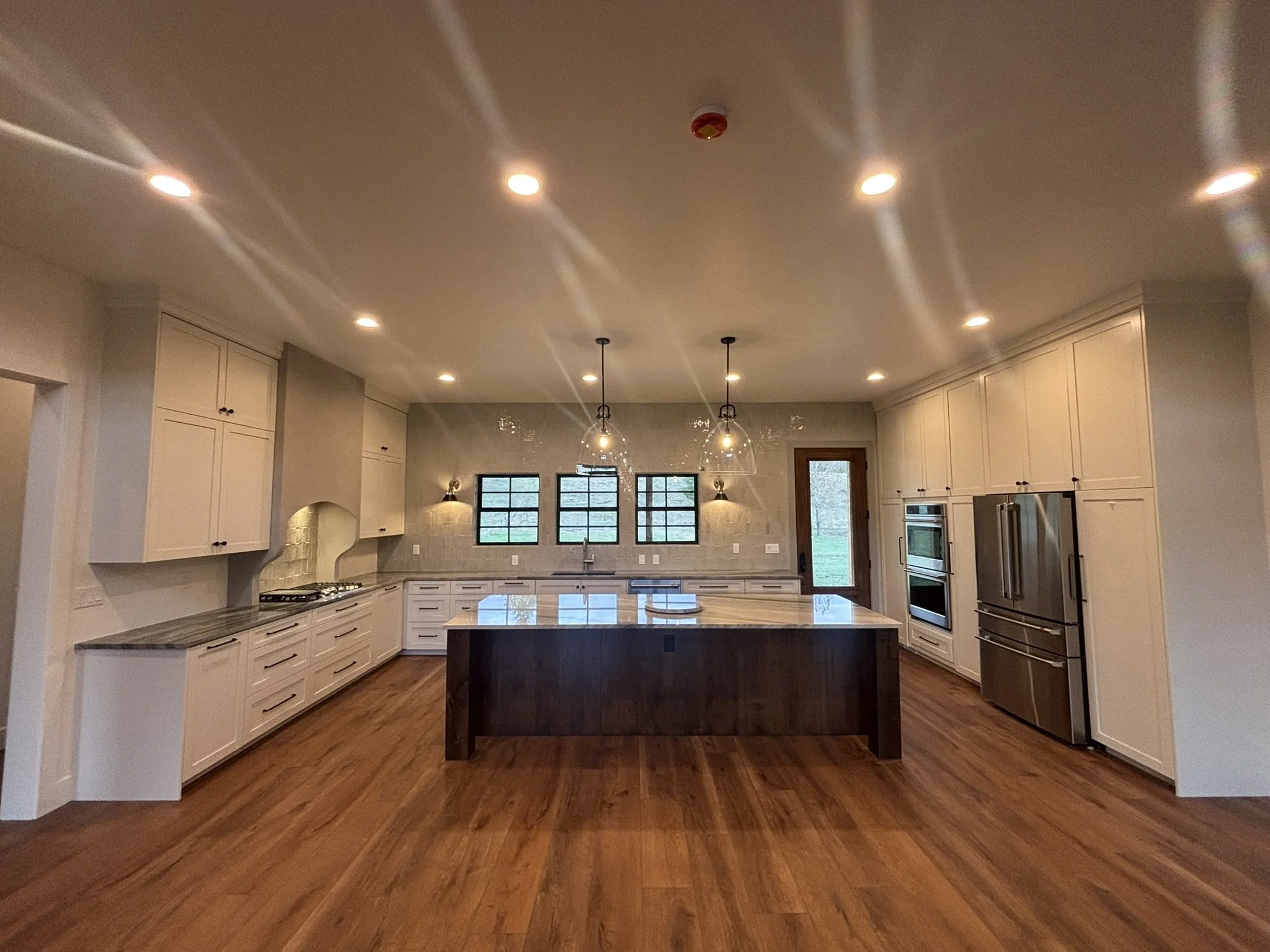 Modern kitchen with a large island, white cabinets, three windows, pendant lighting, and a wooden door.