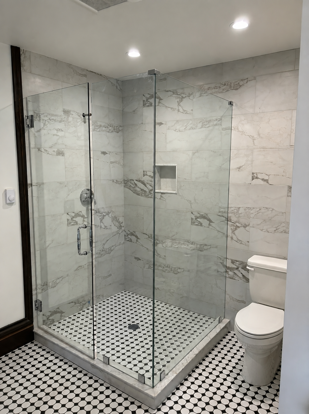 Bathroom with glass shower enclosure and a toilet, featuring marble tiles and a black and white hexagonal tile floor.