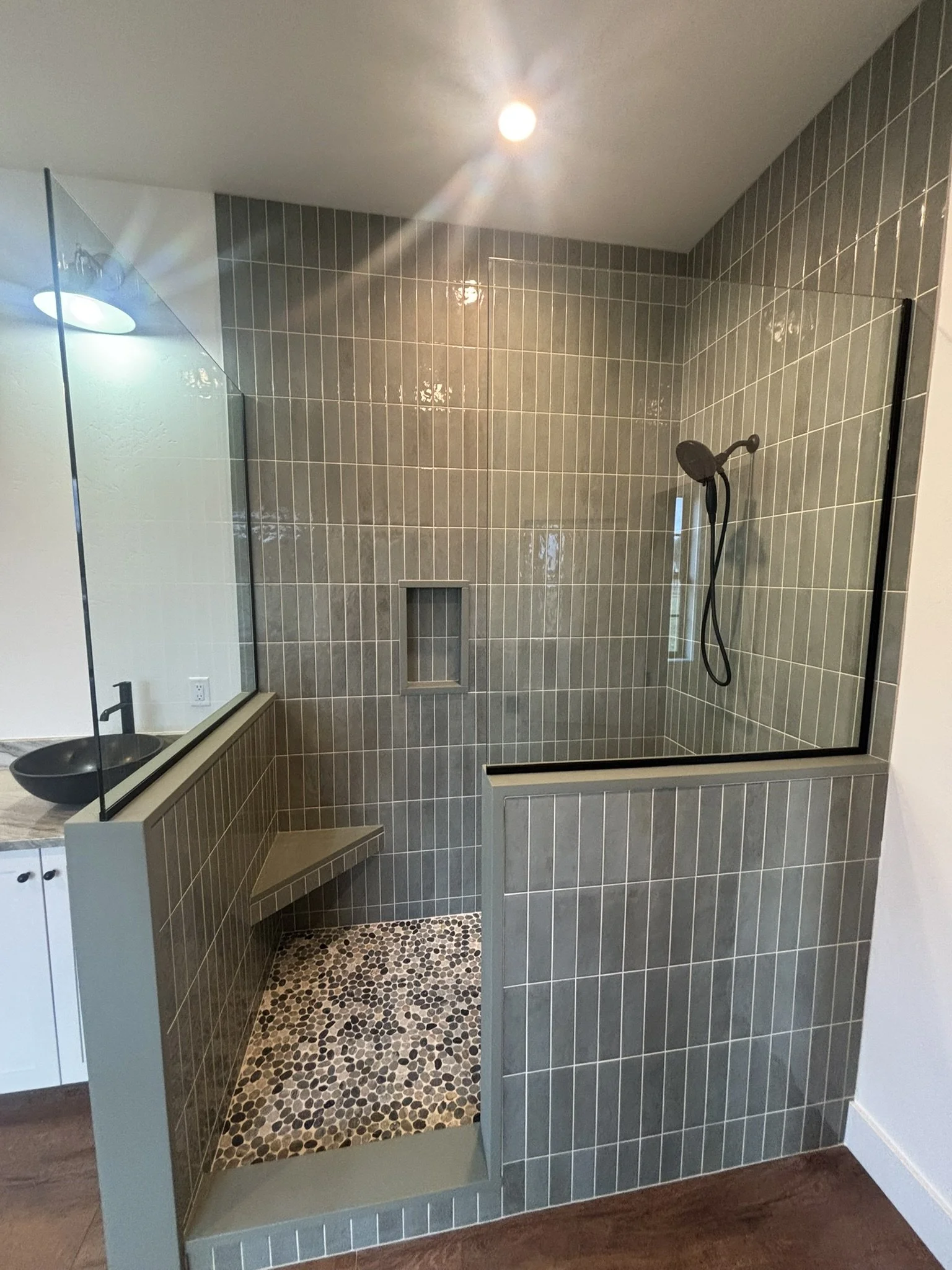 Modern bathroom shower with glass enclosure, gray tiles, pebble floor, and a built-in bench.