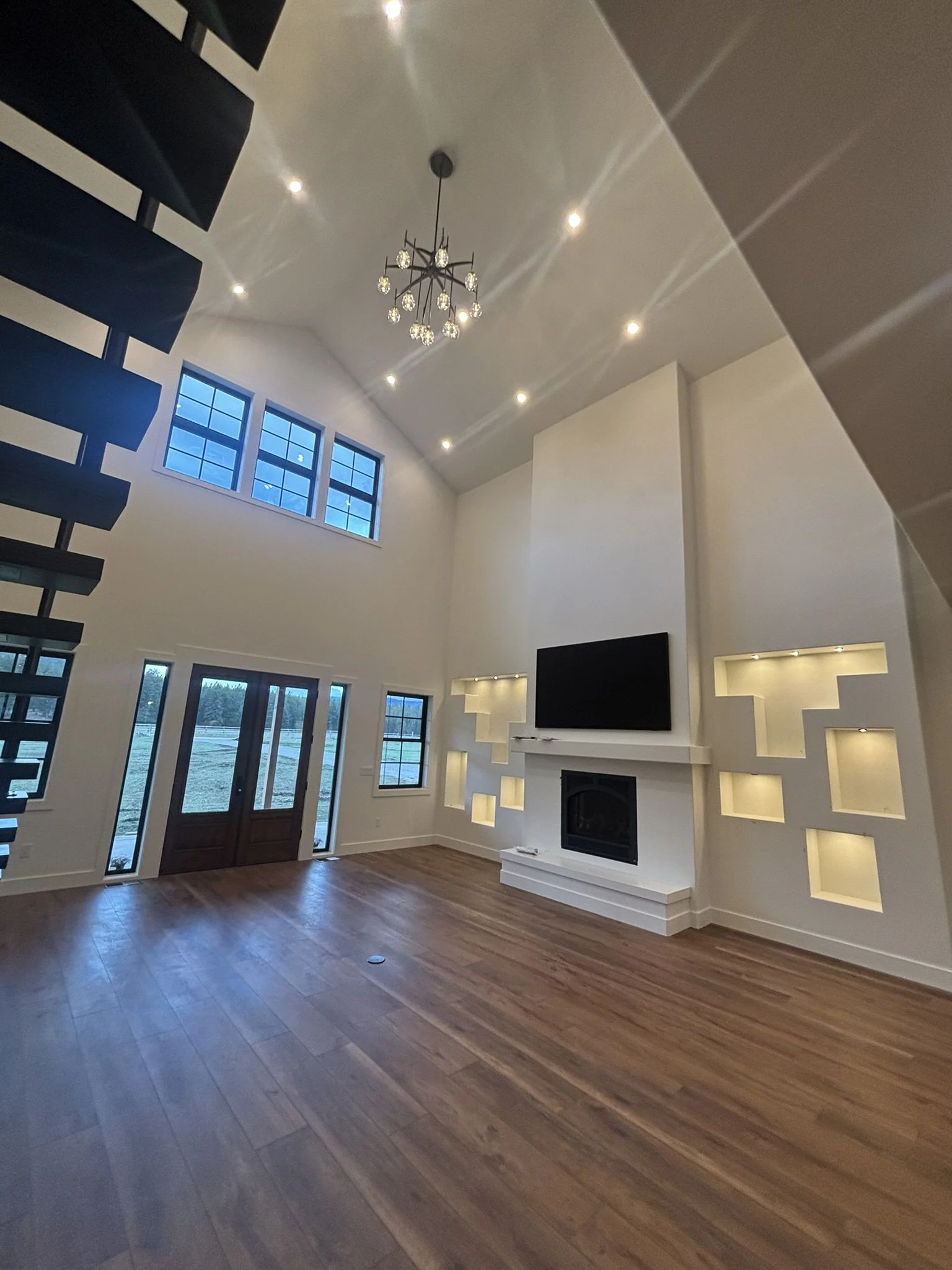 Modern living room with high ceilings, hardwood floors, a contemporary chandelier, a wall-mounted flat-screen TV, a fireplace, and built-in wall niches with lighting.