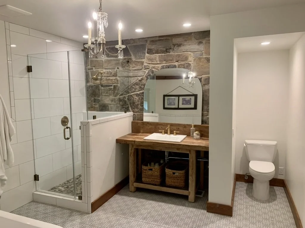 A bathroom with a glass shower, a wooden vanity with a sink, a large mirror, a stone wall, and a chandelier.