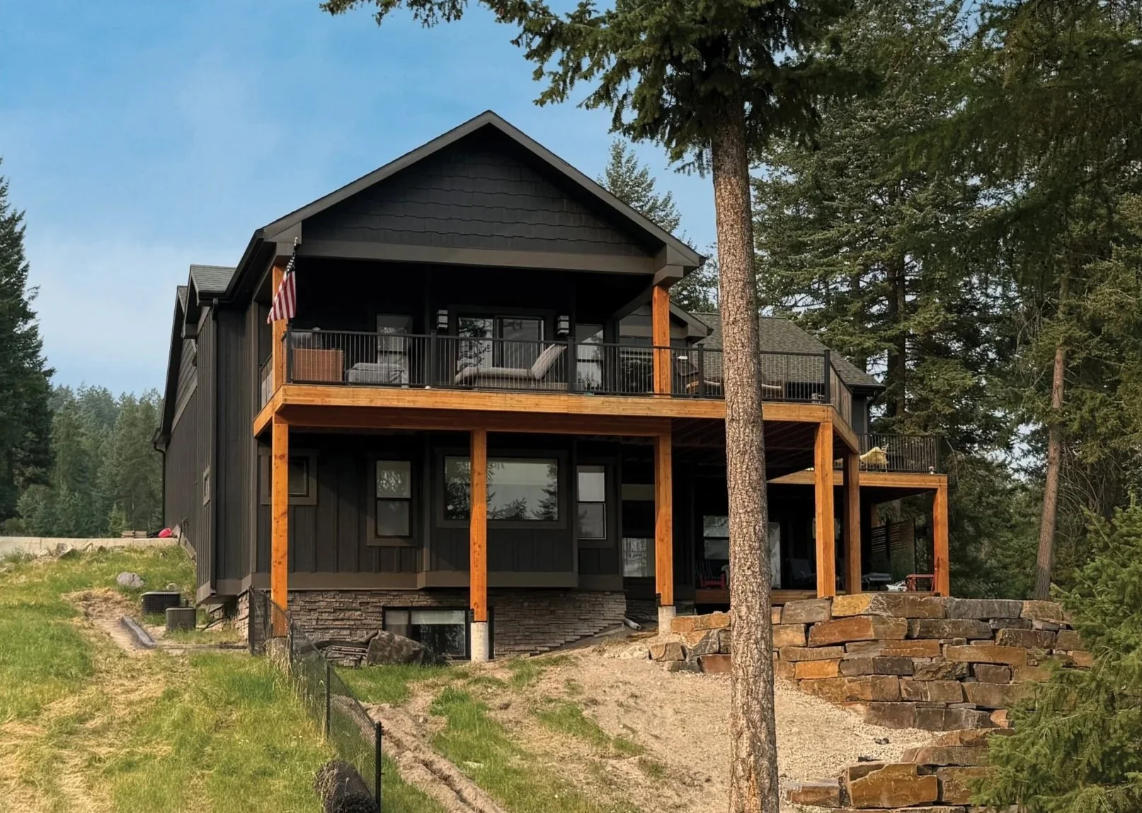 A modern two-story house with outdoor balcony, black siding, surrounded by trees, with a bright blue sky in the background.