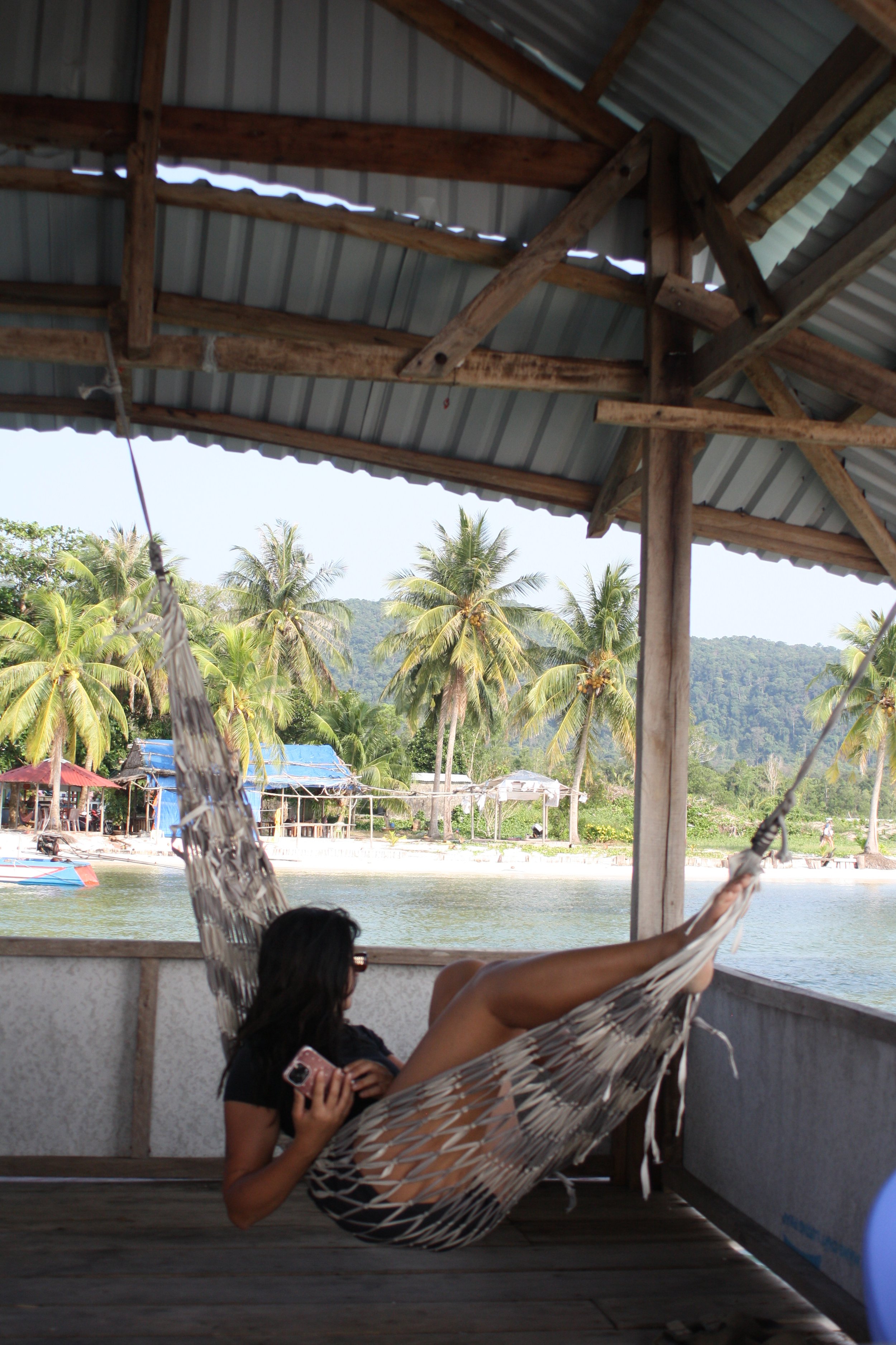 Una mujer acostada en una hamaca en una terraza con vista a un río, árboles de coco y casas en un entorno tropical. isla paradisiaca, tiempo para relax.