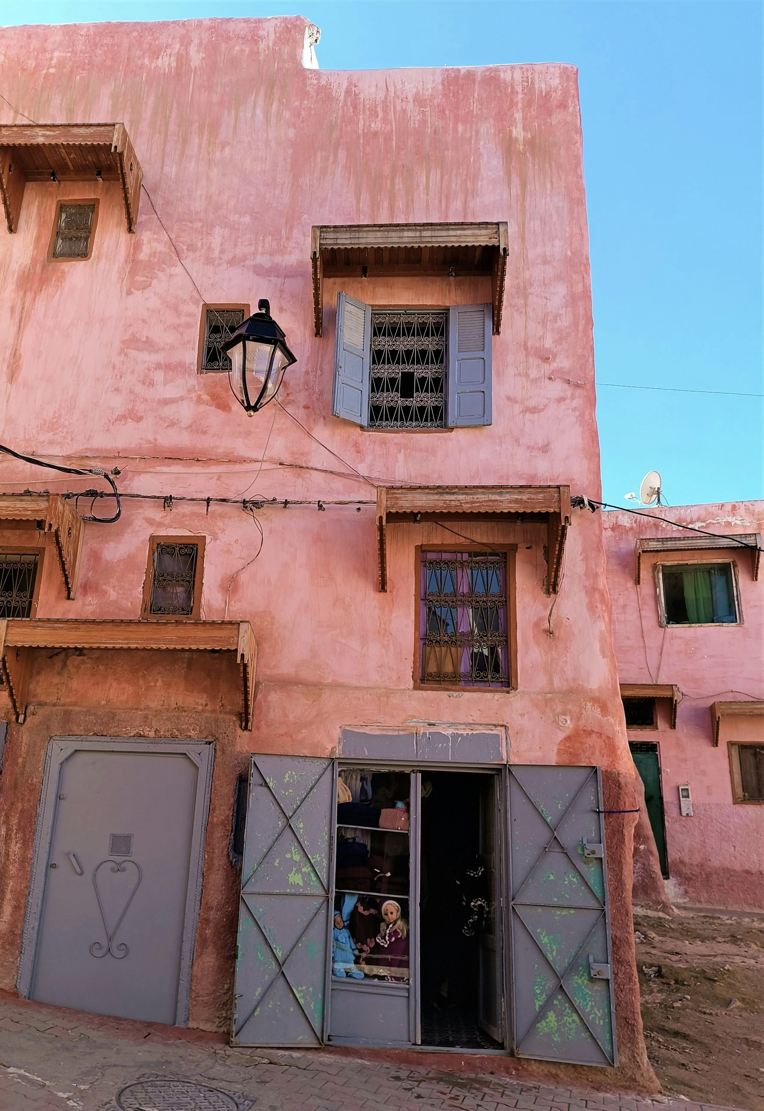 Edificio rosa con ventanas de rejas y balcones de madera, una lámpara de calle negra, cables eléctricos, una puerta metálica de entrada con niños y adultos adentro, y un satélite en la esquina superior derecha.