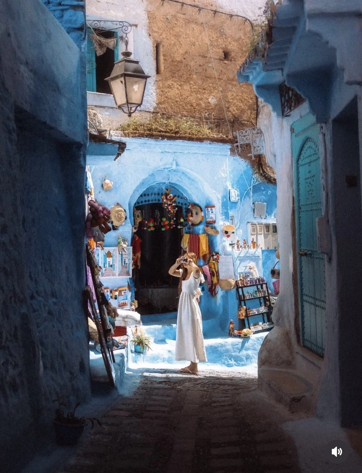 Mujer en vestido blanco tomando una fotografía en una calle artesanal con paredes azules en un entorno típico de ciudad, con puestos de souvenirs y decoraciones coloridas. pueblo azul de marruecos