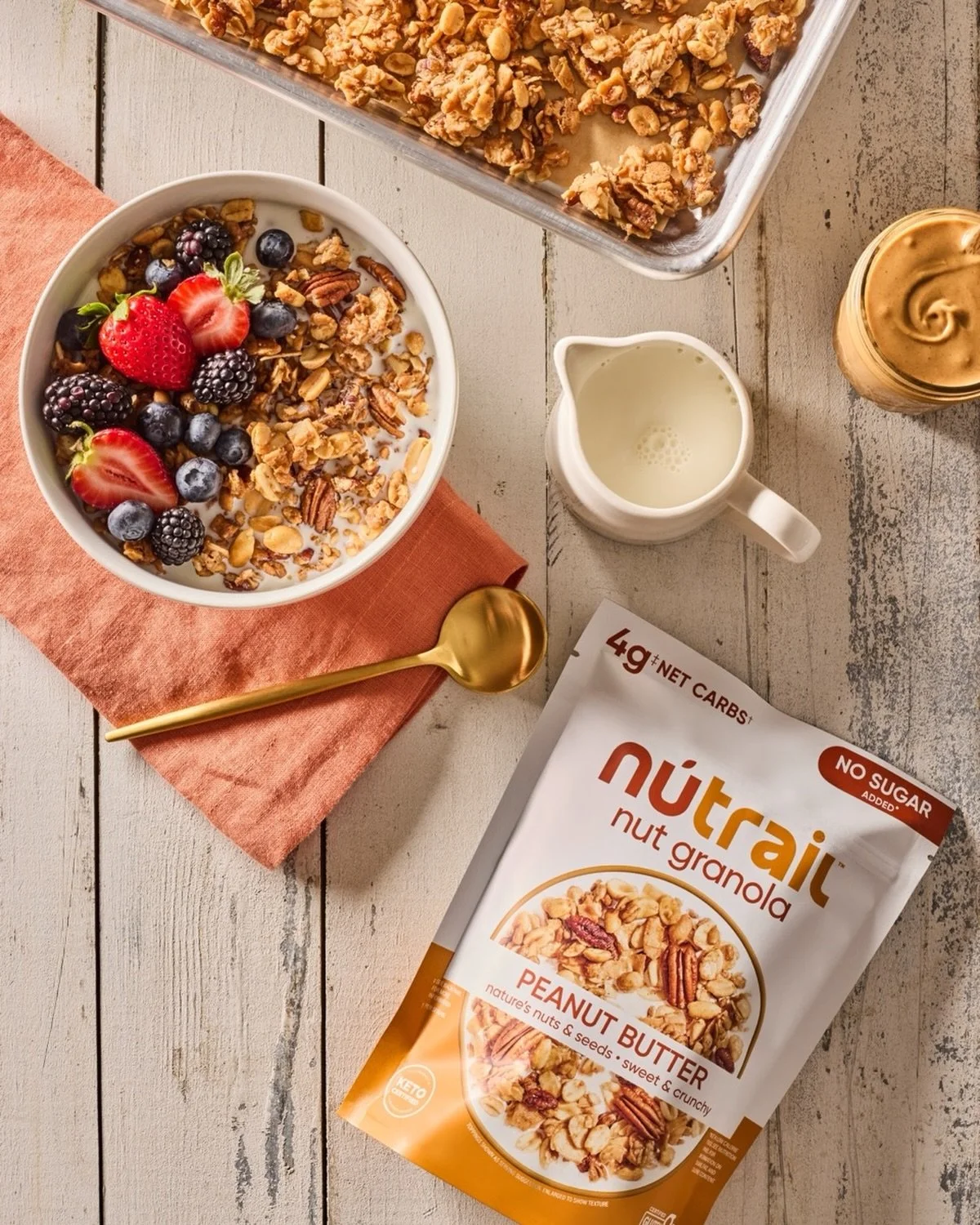 A bowl of yogurt topped with fresh strawberries, blackberries, blueberries, granola, and nuts, placed next to a package of NutraAil peanut butter nut granola, with a jar of peanut butter, a small pitcher of milk, and a golden spoon on a pink napkin, 