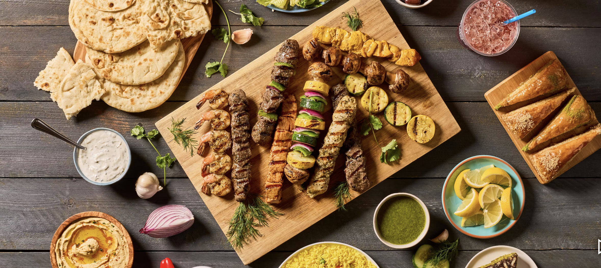 A variety of grilled meats and vegetables on skewers, accompanied by flatbread, sauces, lemon wedges, rice, and drinks on a wooden table.