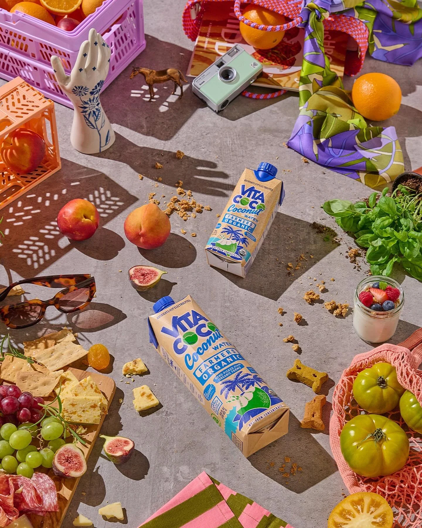 A picnic scene with various food and items scattered on the ground. There are apples, grapes, figs, cheese, crackers, a jar of yogurt with berries, cartons of coconut water, a small toy horse, sunglasses, a camera, and colorful fabric and baskets. Th