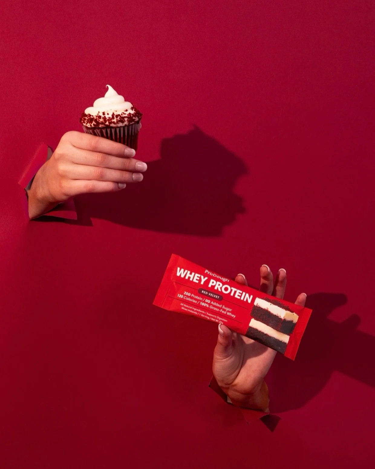 A person's hand holding a package of whey protein with a red background. The other hand is holding a cupcake with white frosting and red sprinkles.