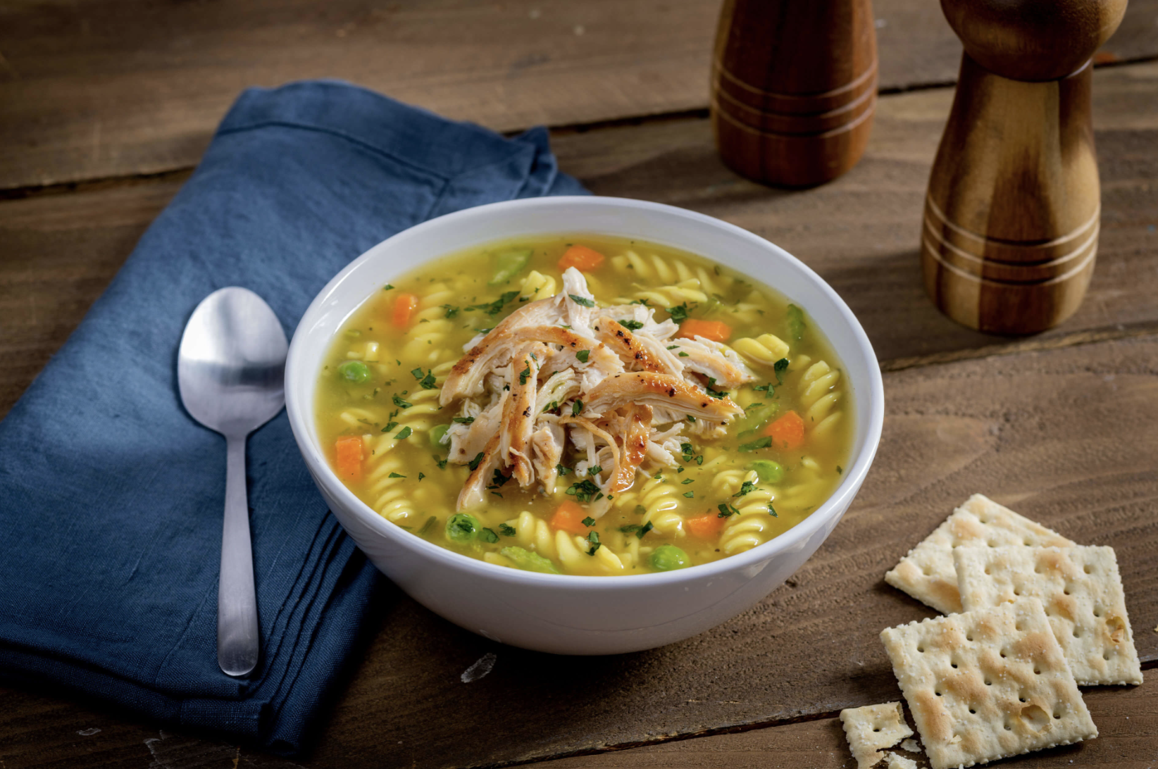 Bowl of chicken noodle soup with shredded chicken, carrots, peas, and herbs on a wooden table. A blue napkin with a spoon, salt and pepper shakers, and saltine crackers nearby.