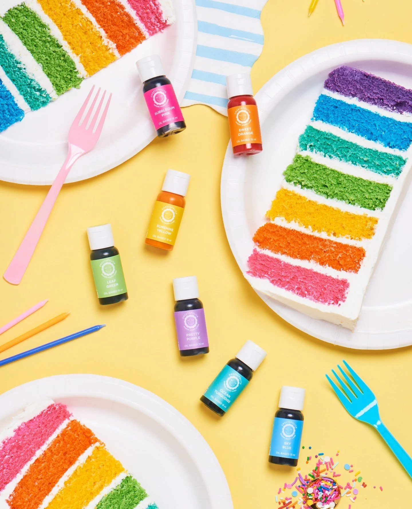 Colorful rainbow cake slices on white plates, rainbow sprinkles, and bottles of colored food coloring on a yellow background, with plastic forks and a cupcake with sprinkles.