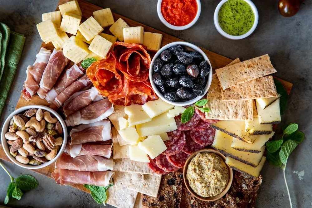 Charcuterie board with cheeses, cured meats, black olives, and nuts, accompanied by small bowls of red and green sauces.