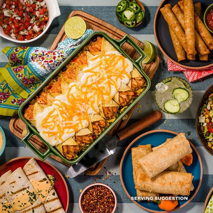 A variety of Mexican dishes including a casserole topped with cheese and sour cream, fried spring rolls, quesadillas, sliced cucumber, salsa, and chopped tomatoes, served with lime wedges.