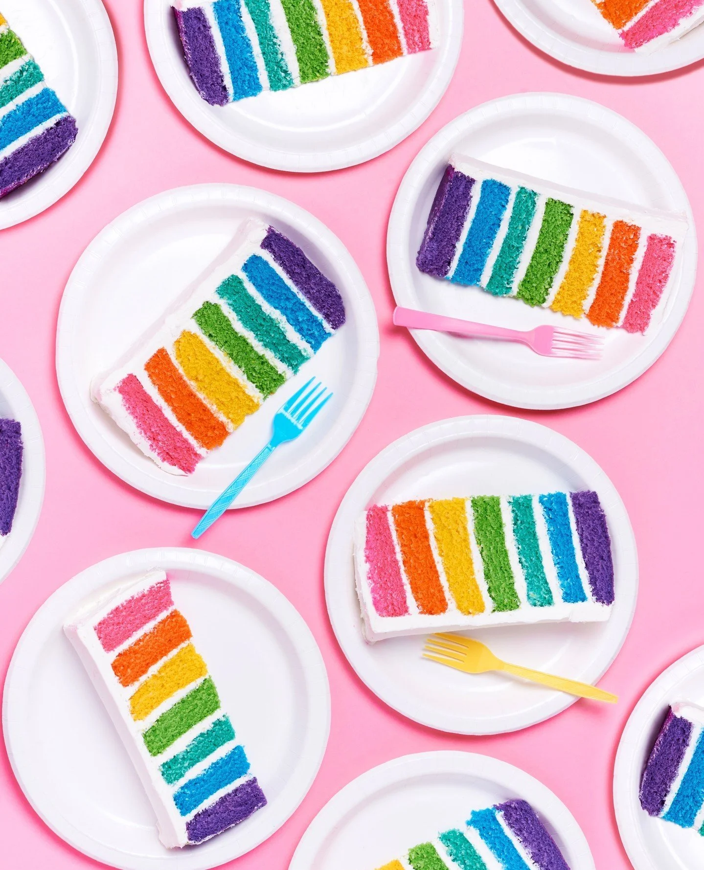 Multiple slices of rainbow layered cake with bright colors on white paper plates, set against a pink background, with pastel-colored forks.