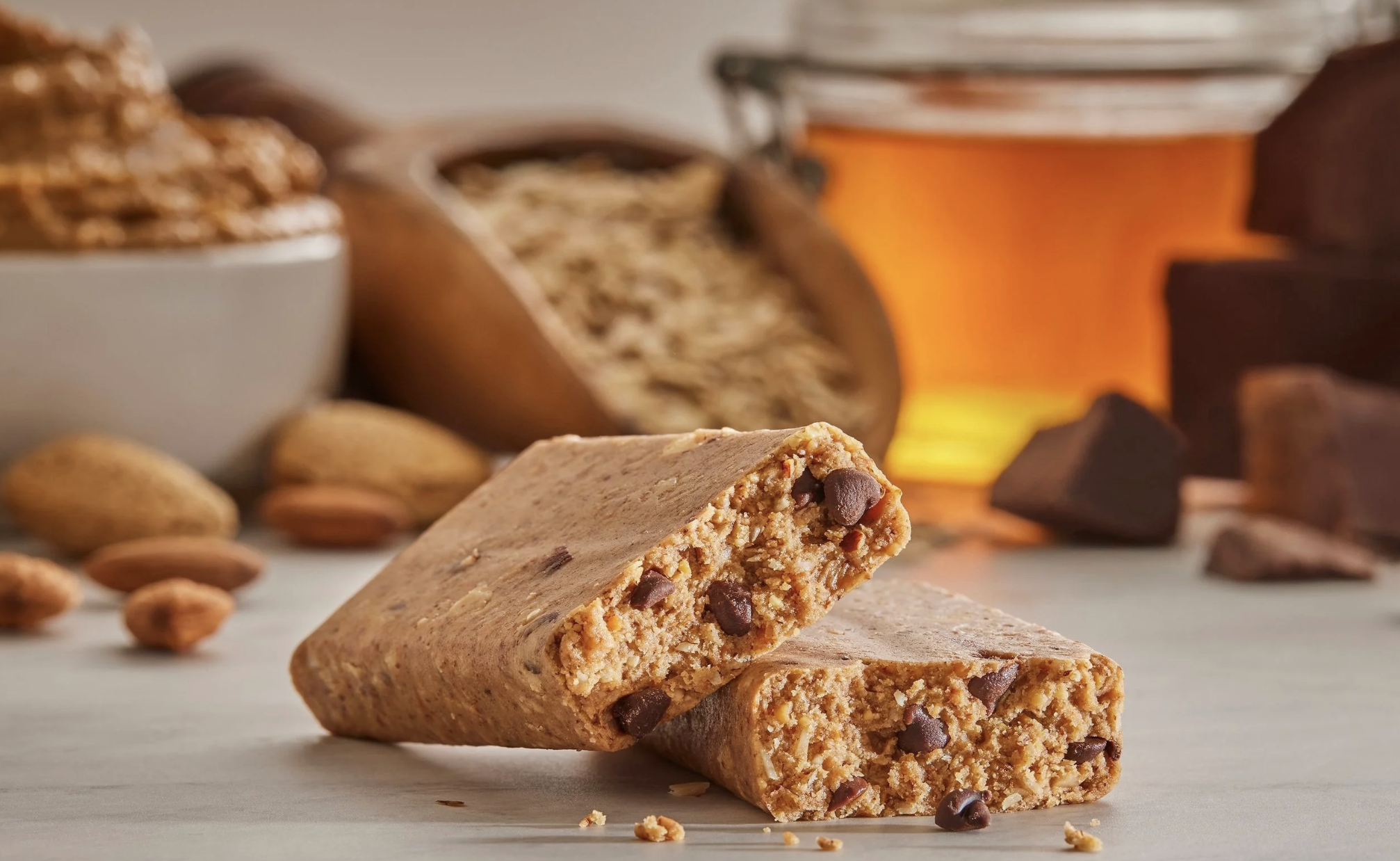 Close-up of two almond and chocolate chip cookie bars on a light-colored surface surrounded by whole almonds and chocolate pieces, with blurred background of a bowl of almonds and a jar of honey.