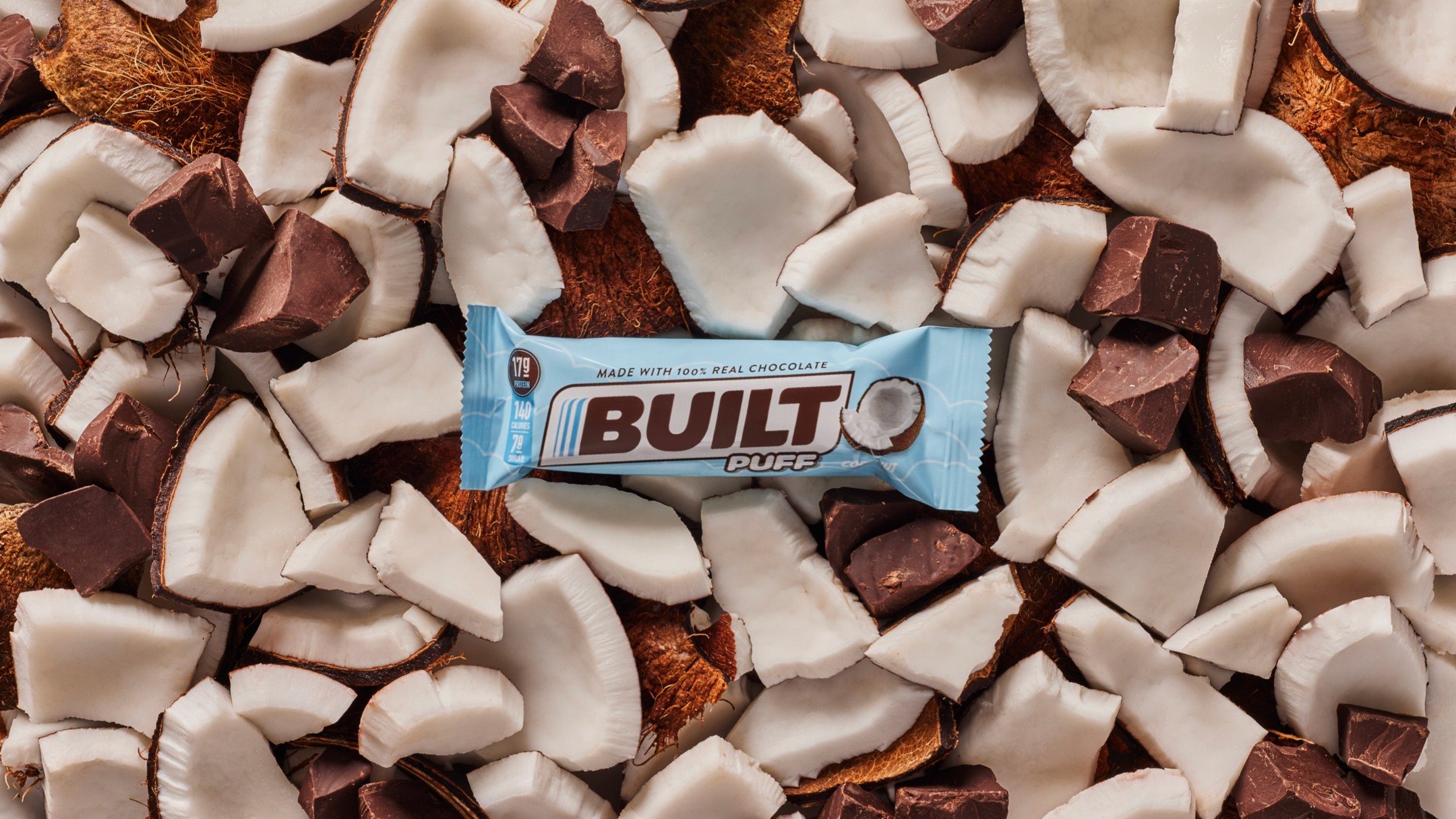 A close-up image of chopped coconut pieces and chunks of chocolate on a wooden surface with a mint blue Built Puff wrapper in the center.