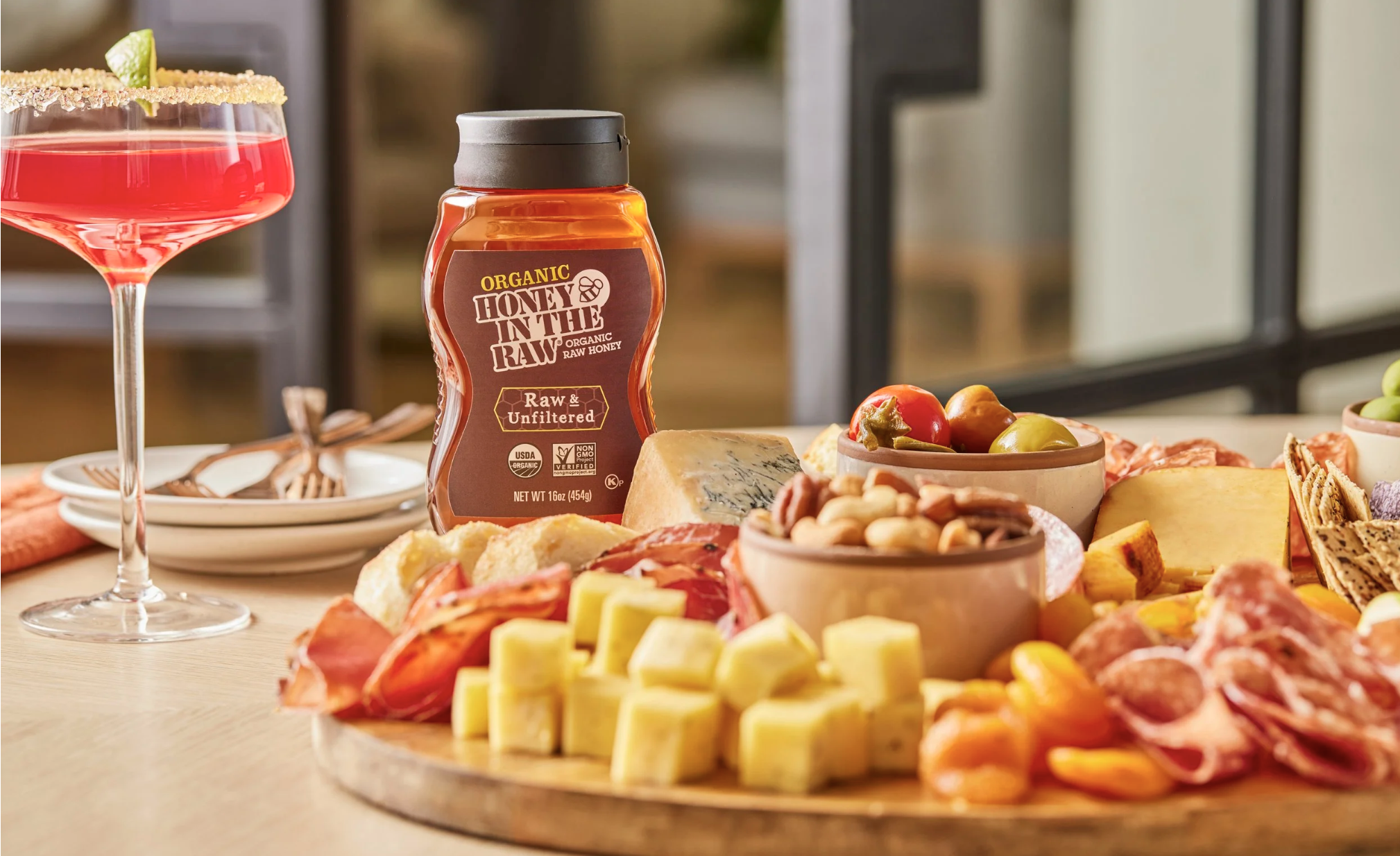 A table filled with cheese, meats, nuts, fruits, and vegetables, with a cup of honey in the center and a martini glass with a lime garnish.