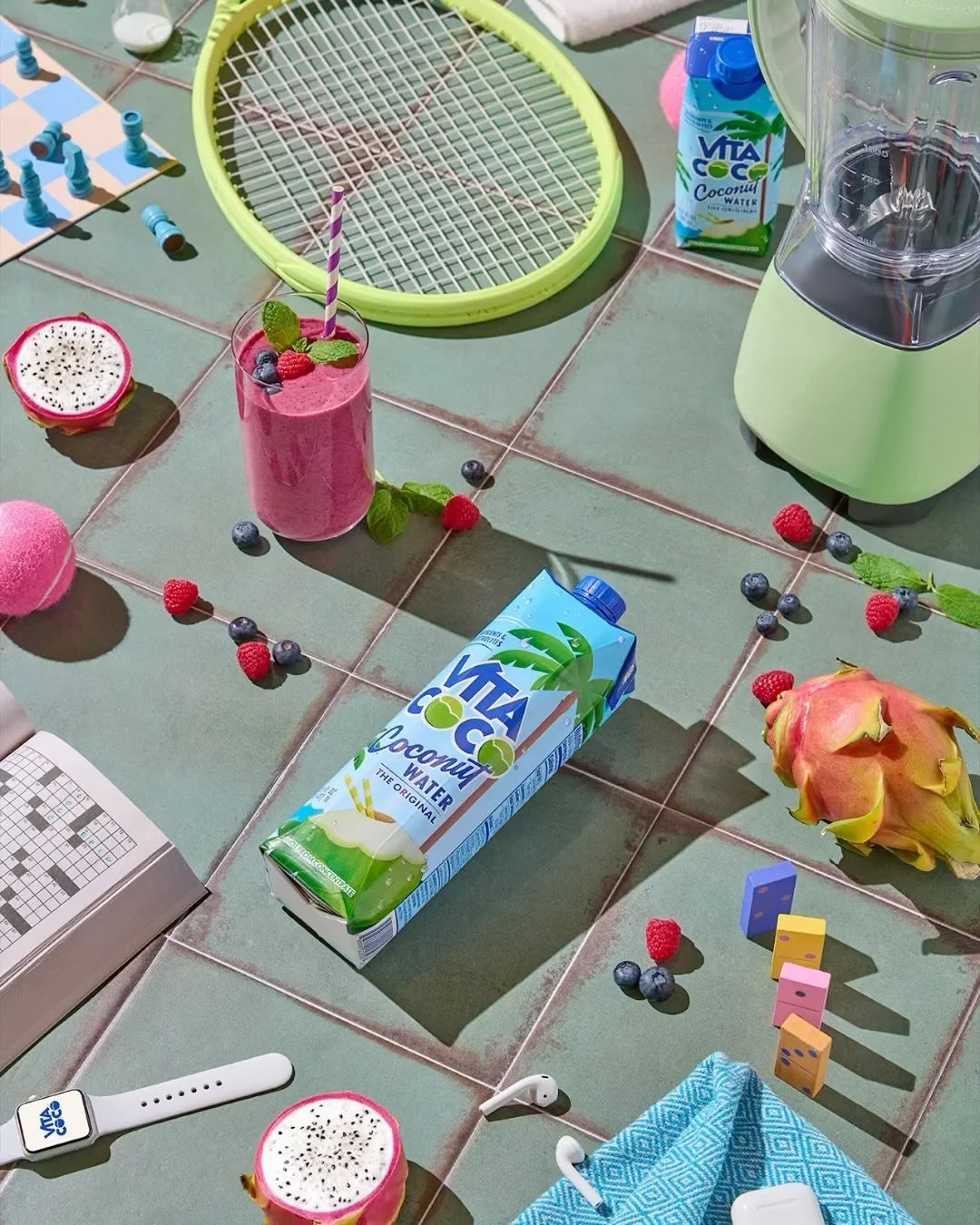 Tiled outdoor table with a pink smoothie topped with berries and mint, a carton of coconut water, a dragon fruit, scattered berries and mint leaves, an alarm clock, a pair of in-ear earbuds, a blue cloth, domino pieces, a tennis racket, and a blender