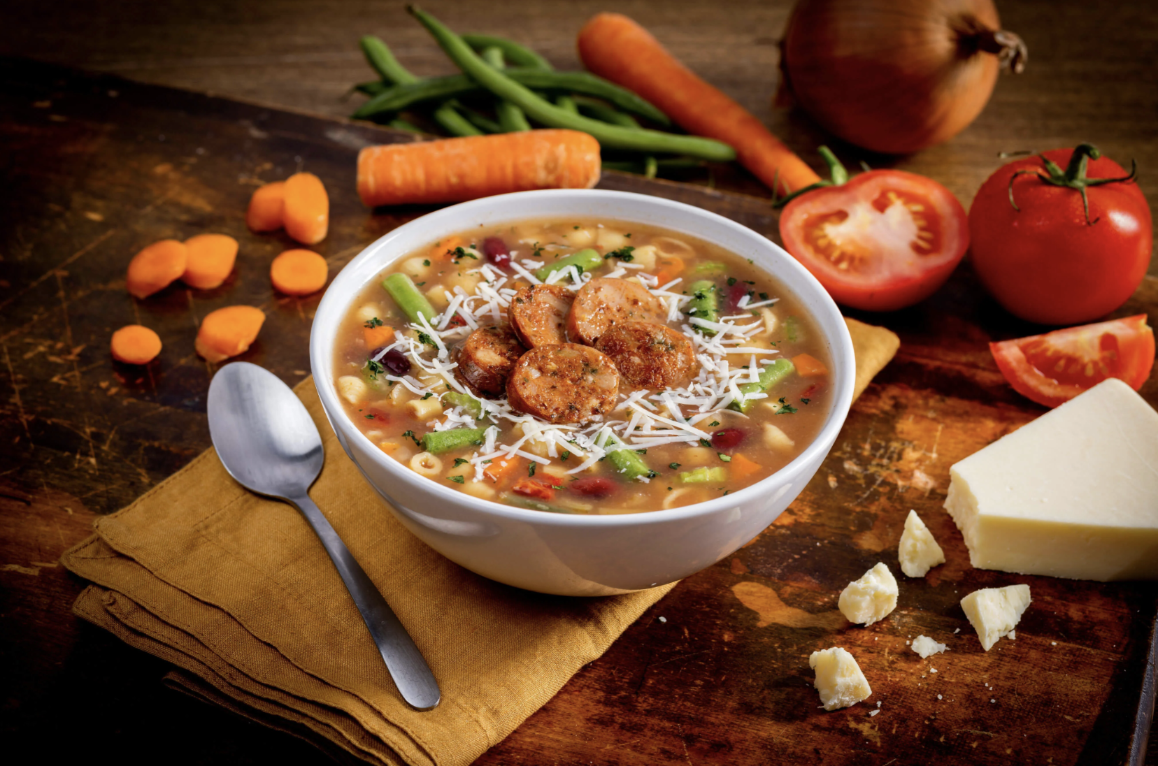 A bowl of chicken noodle soup topped with shredded cheese and sliced sausage, on a wooden table surrounded by vegetables, cheese, and a spoon.