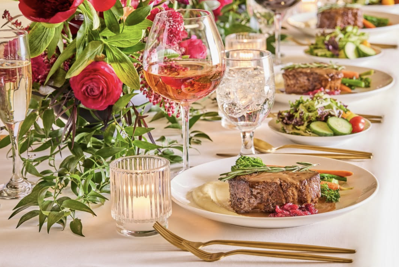Elegant dining table set with plates of steak and vegetables, glasses of rosé wine and water, candlelight, and a floral centerpiece with red and pink flowers.