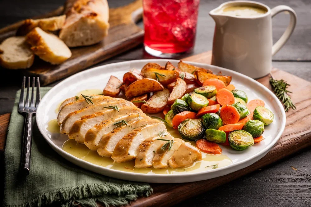 Cooked sliced chicken breast with herbs, roasted potatoes with herbs, and cooked carrots and Brussels sprouts on a white plate, with a glass of red beverage, a bowl of gravy, and bread in the background.