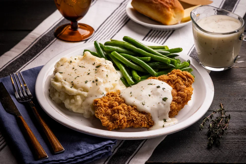 Fried chicken with white gravy, mashed potatoes with gravy, green beans, a dinner roll, and a glass of iced tea on a dark wooden table.