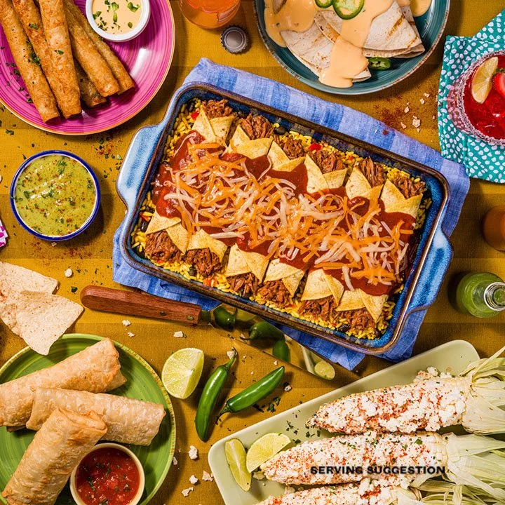 A Mexican meal table with a tray of tacos, a casserole of seasoned rice topped with shredded cheese, nachos, spring rolls with salsa, and a bowl of guacamole with chips.
