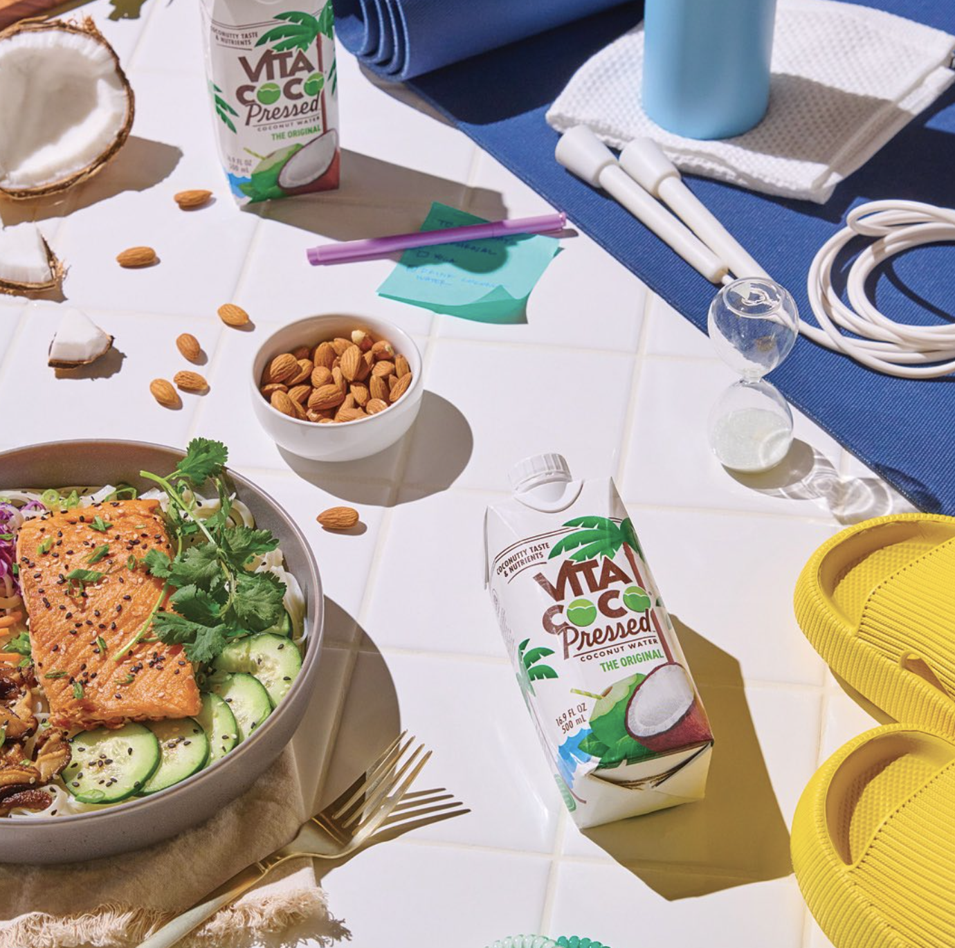 A table with a bowl of salad, a carton of Vita Coco coconut water, a small bowl of almonds, and various beach items including yellow flip-flops, a snorkel mask, and a wrapped towel.