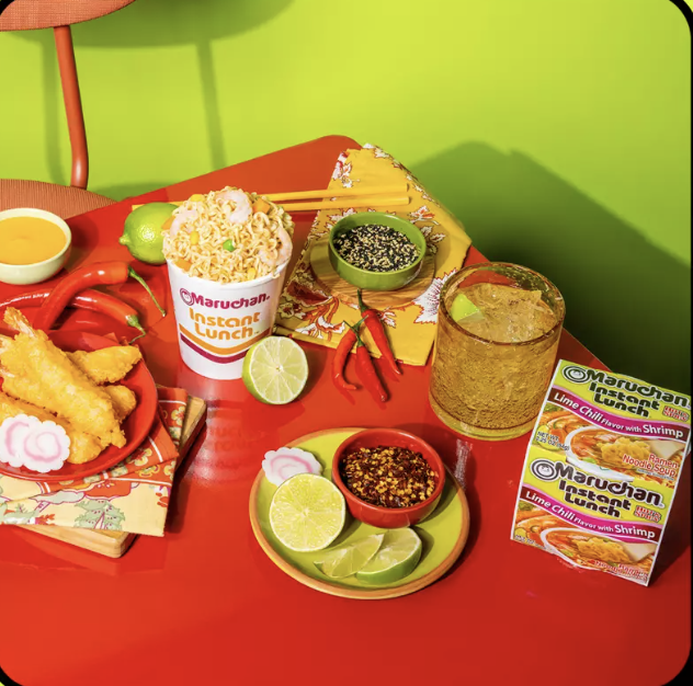 A red table with various Asian-themed food and drink items, including instant noodles, lime wedges, chili peppers, dipping sauces, tempura, and a glass of a brown beverage, against a green wall background.