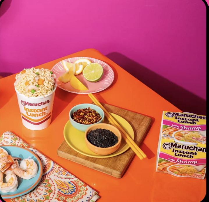 A colorful table setting with Maruchan Instant Lunch cups, a small plate of lemon slices, a bamboo cutting board with soy sauce and black pepper, a bowl of red pepper flakes, a bowl of black caviar, and a plate of cooked shrimp on a decorative napkin