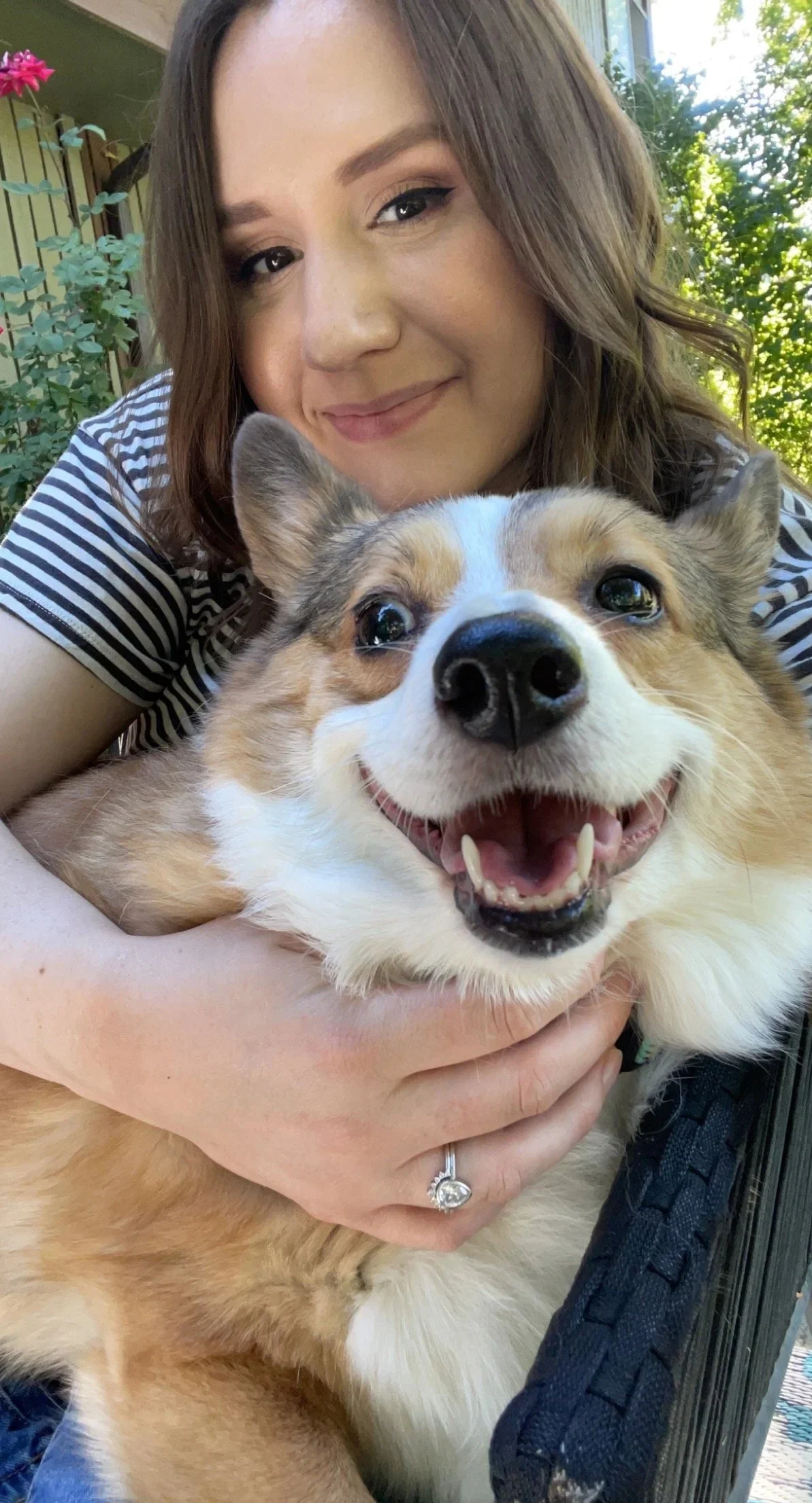 A woman with light brown hair smiling and holding a smiling, fluffy, tan and white dog outdoors on a sunny day.