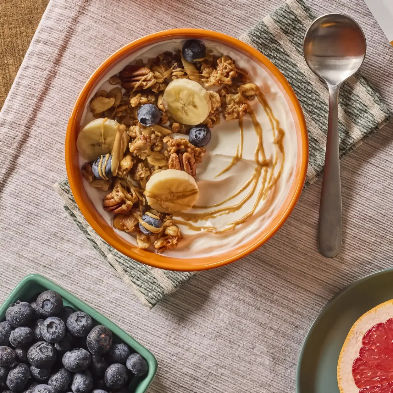 A bowl of yogurt topped with granola, sliced bananas, blueberries, and drizzled with honey, placed on a striped cloth with a scoop next to it and a green bowl of blueberries at the bottom left.