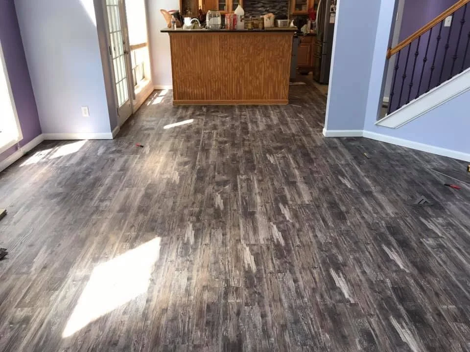 Living room with dark textured wood flooring, light purple walls, and a kitchen in the background with a wooden counter. There's a staircase with dark handrails on the right, and sunlight coming through windows on the left.