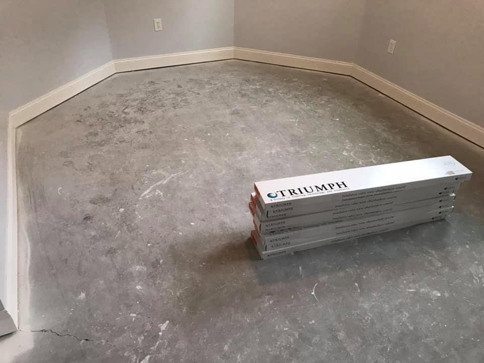 Empty room with gray walls and baseboards, concrete floor, with a stack of four boxes labeled 'TRIUMPH' placed on the floor.