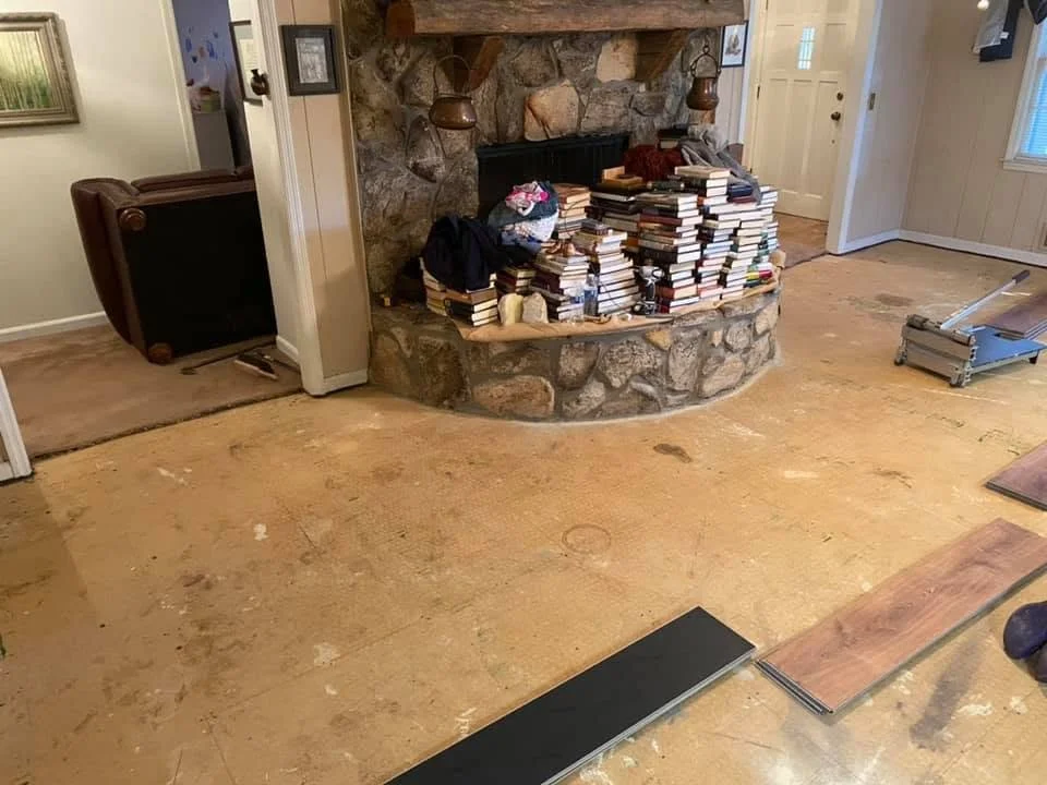 Living room with a stone fireplace, piles of books on the hearth, and a partially finished wooden floor.