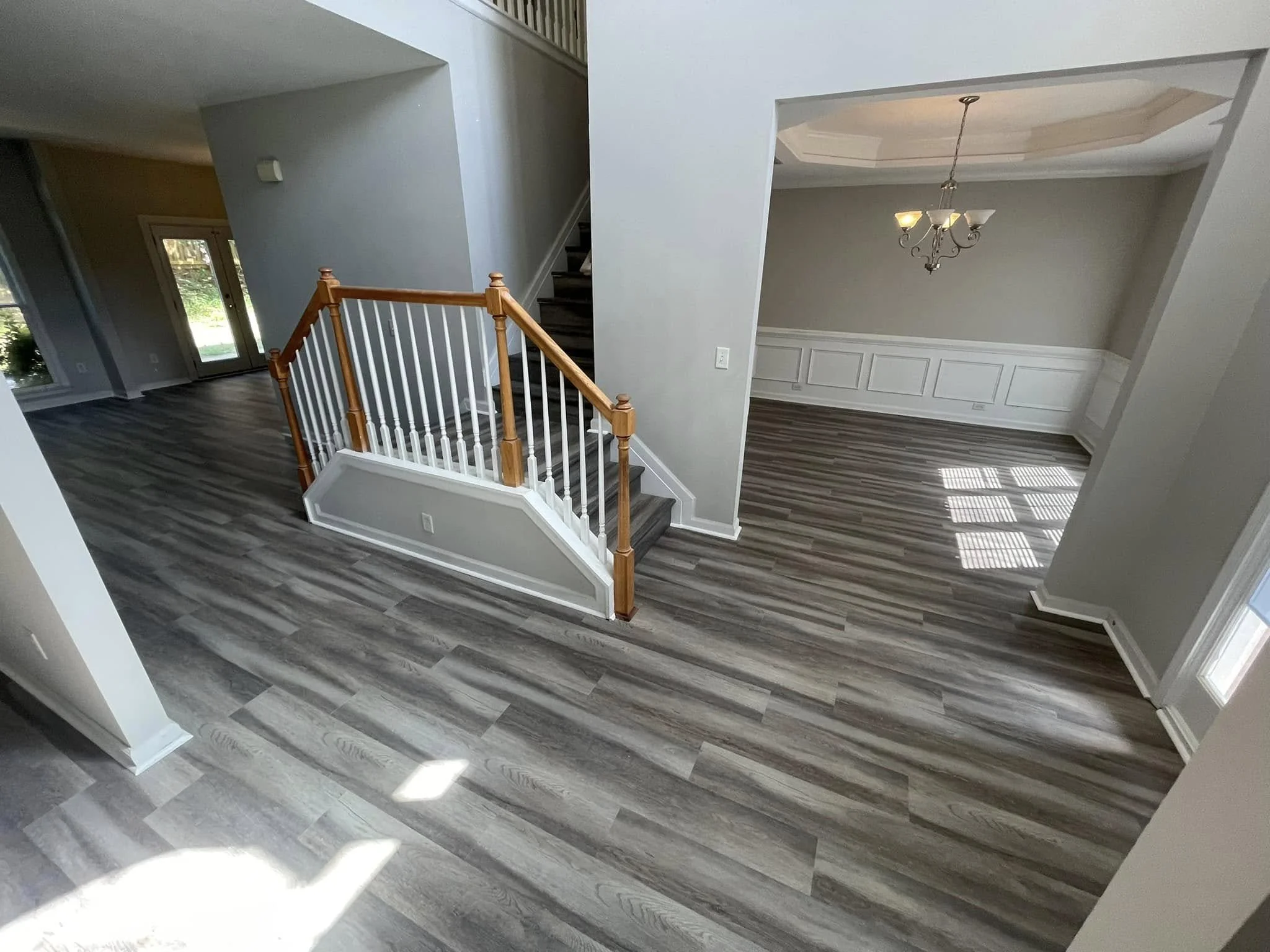 Interior view of a modern home with hardwood floors, staircase with wooden railing, large window and a hanging chandelier in the dining area.