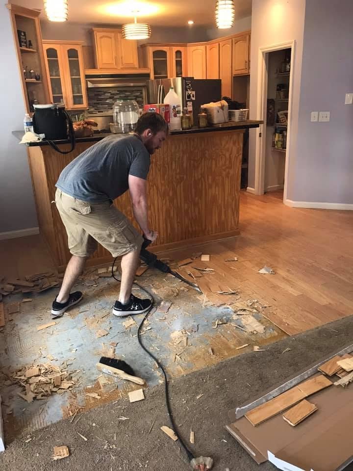 A man is removing old wood flooring with a crowbar and a sledgehammer in a kitchen area, with scattered wood debris on the floor.