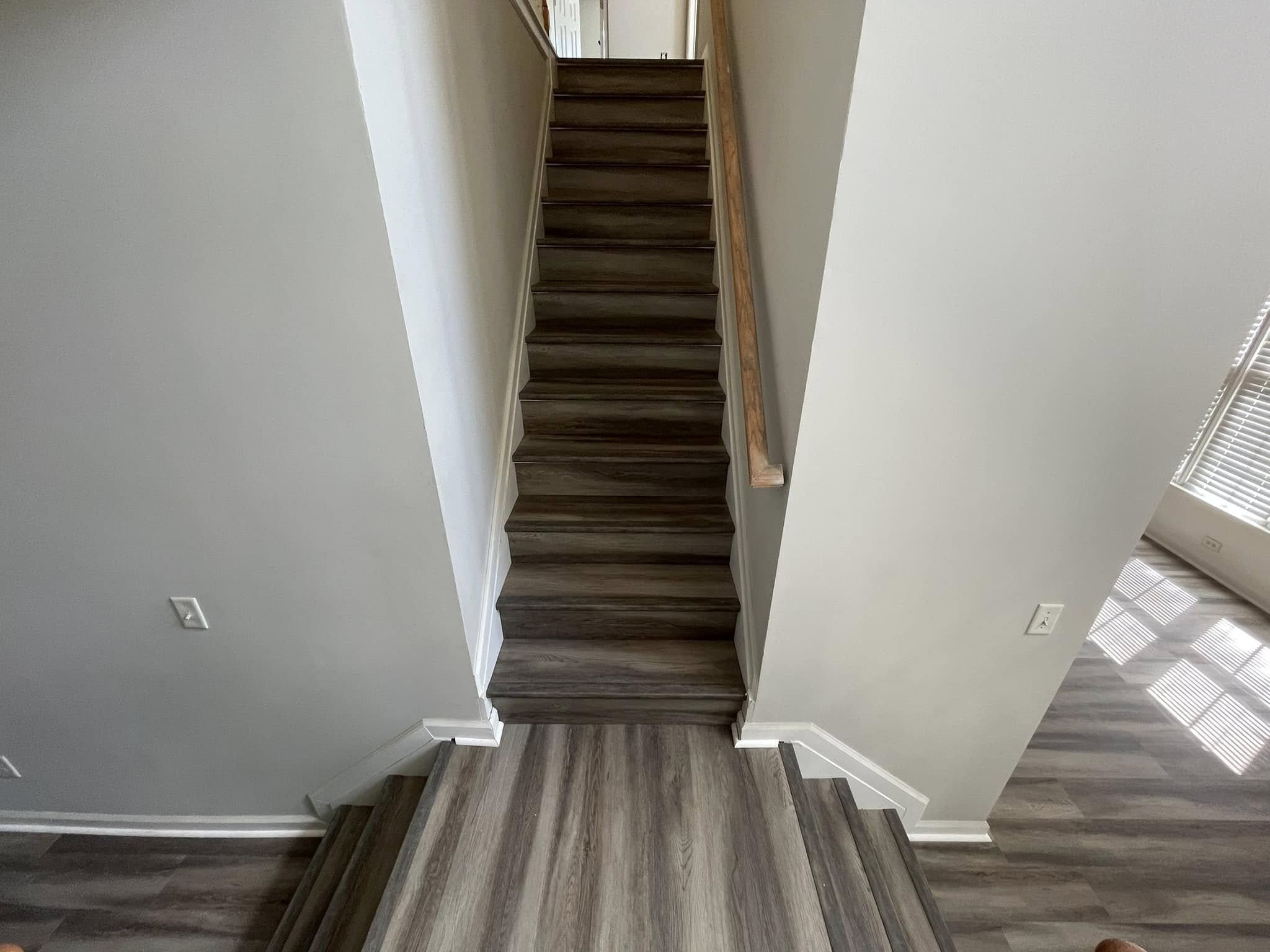 Interior view of a staircase with dark wooden steps, beige walls, white baseboards, and hardwood flooring, with sunlight coming through a nearby window.