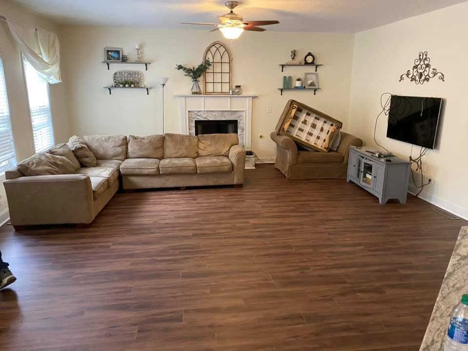 Living room with beige sectional sofa, fireplace, wall-mounted TV, wooden floor, ceiling fan, and decorative wall hangings.