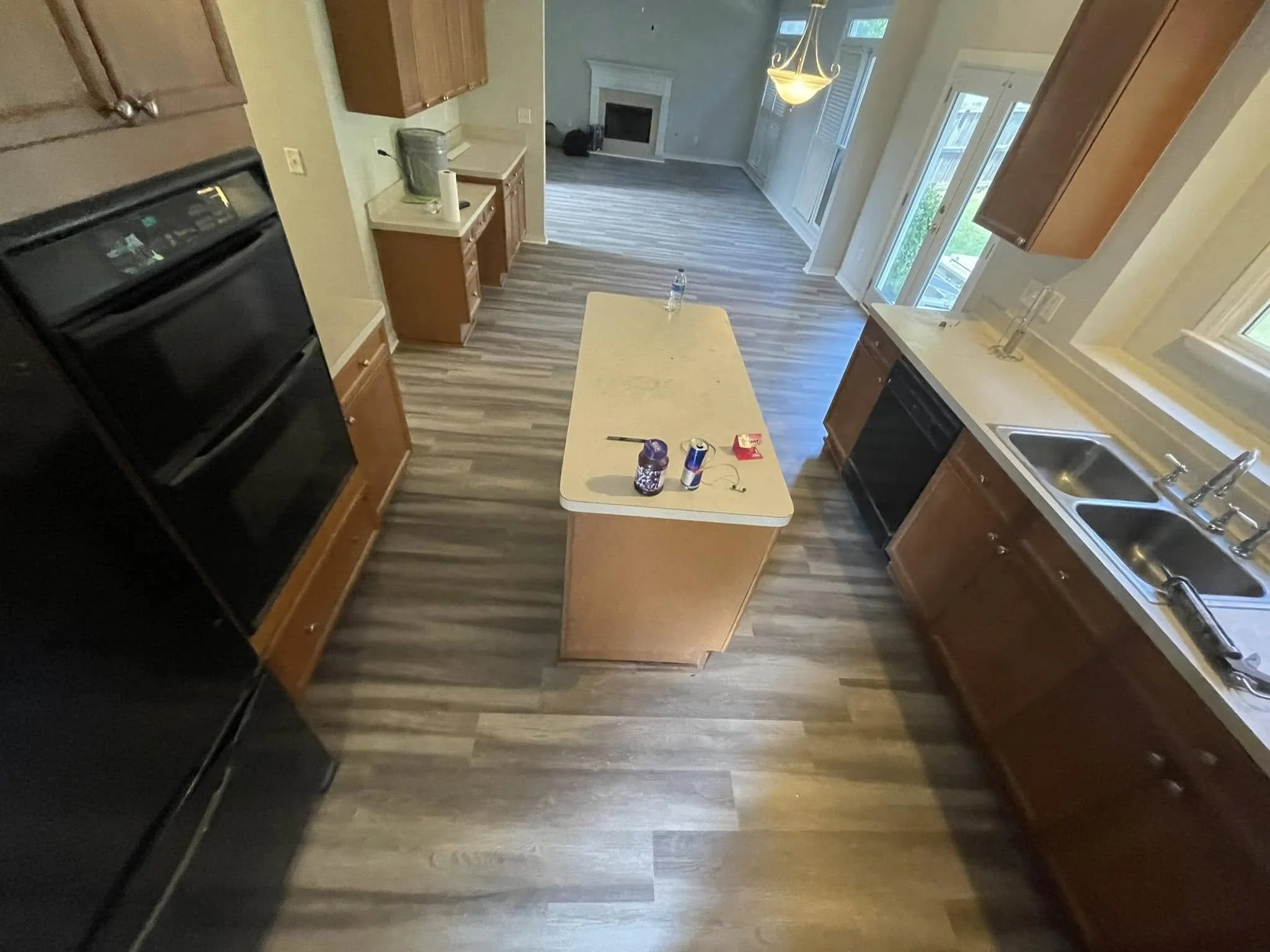 View of a kitchen with wooden cabinets, a beige countertop, black appliances, a small table with items on it, and a living room area with a fireplace, large windows, and a hanging light fixture.