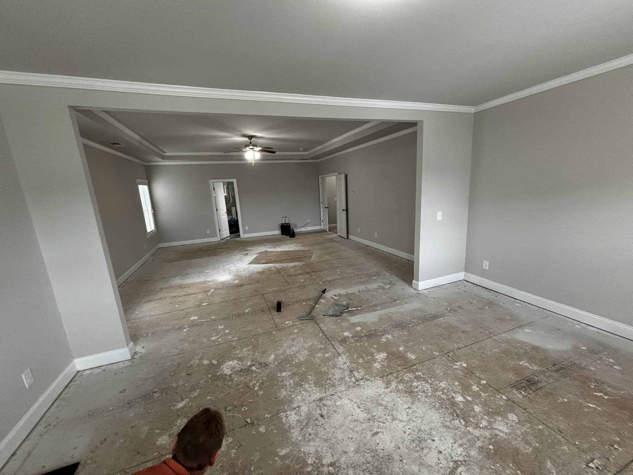 Empty living room with unfinished flooring, light gray walls, white crown molding, ceiling fan, and two windows. Small items on the floor and in the corner.