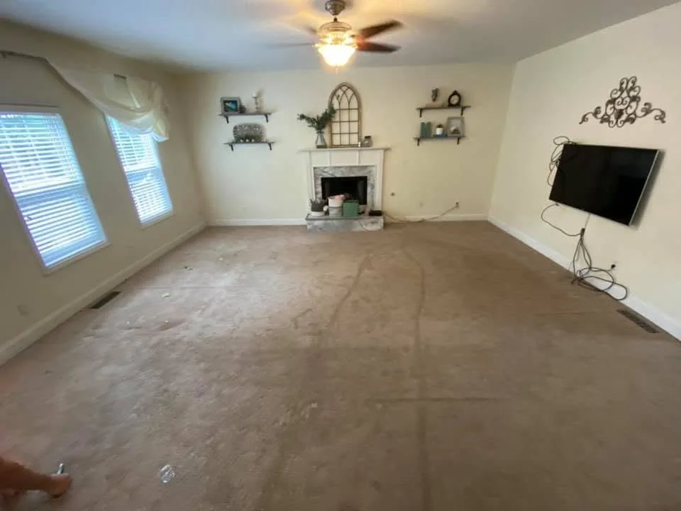 Empty living room with beige carpet, three windows with white blinds and curtains, a ceiling fan with light, a mounted flat-screen TV, a fireplace with decorative items, and wall-mounted shelves with decor.