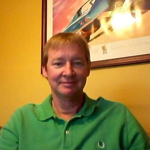 A man with light brown hair and a slight smile, wearing a green polo shirt with a small logo, sitting indoors against a yellow wall with a framed poster behind him.