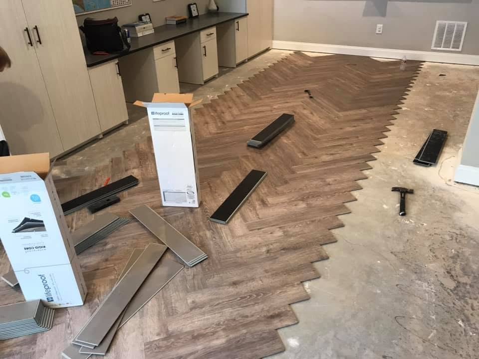A room with hardwood flooring being installed in a herringbone pattern. Various flooring planks, tools, and packaging are visible, with some planks already laid and others waiting to be installed.