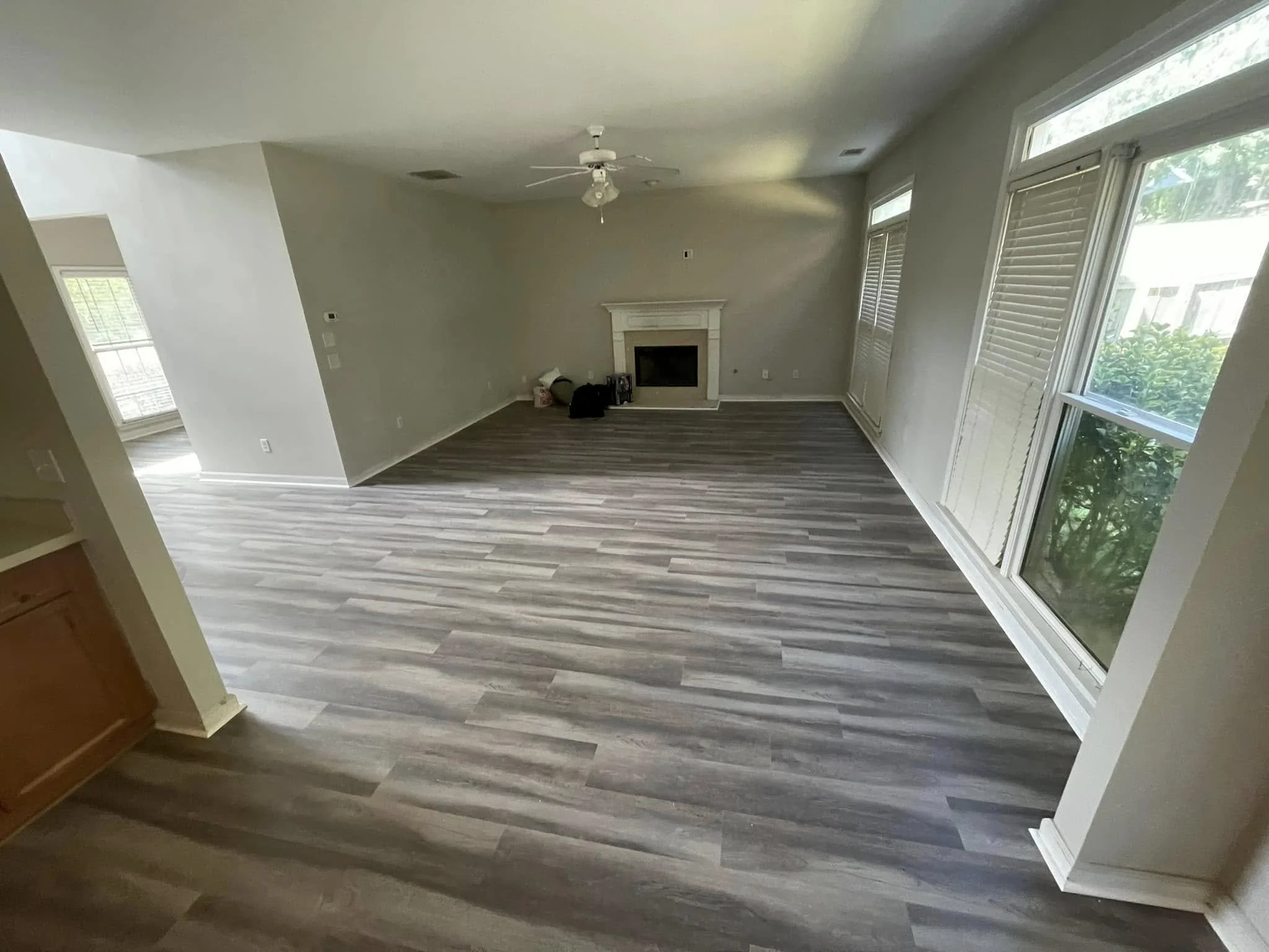 Living room with large windows, fireplace, ceiling fan, and wood floor, with some bags and objects near the fireplace.